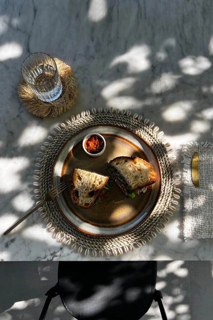 Broodje geserveerd op een placemat van zeegras en kauri schelpen, naast een glas water, op een marmeren tafel.