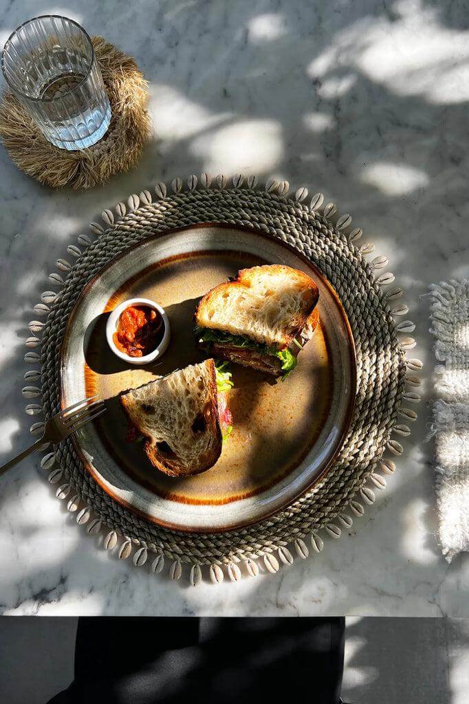 Gezellige tafelsetting met placemat van zeegras en schelpen, een sandwich op een bord en een glasje water. Tropische sfeermaker!