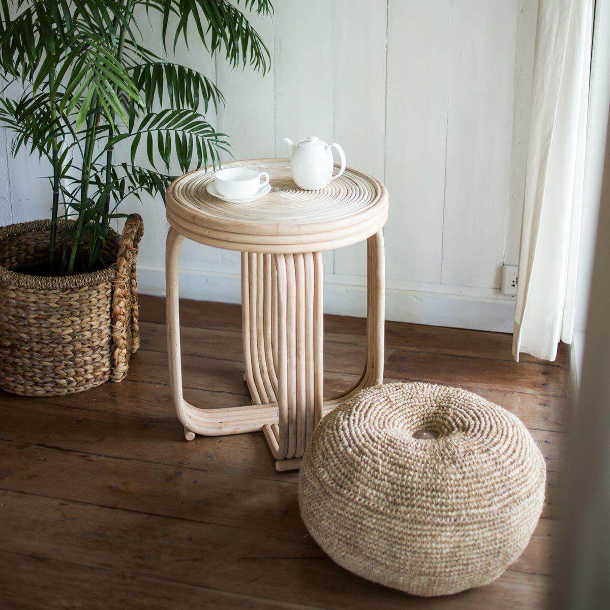 Raffia poef Telu naast houten tafel in woonkamer, eco-vriendelijk en handgemaakt, perfect voor extra zitplaats of voetenbankje.