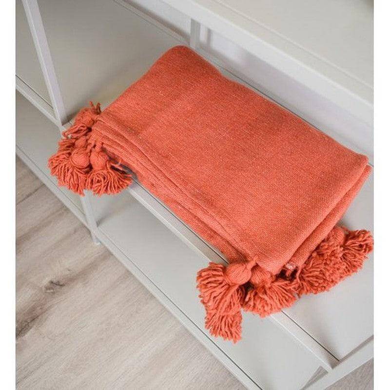 Oranje Marokkaanse pompomdeken op een witte plank. Perfect voor een knusse sfeer in de slaapkamer of woonkamer. Stijlvol en veelzijdig.