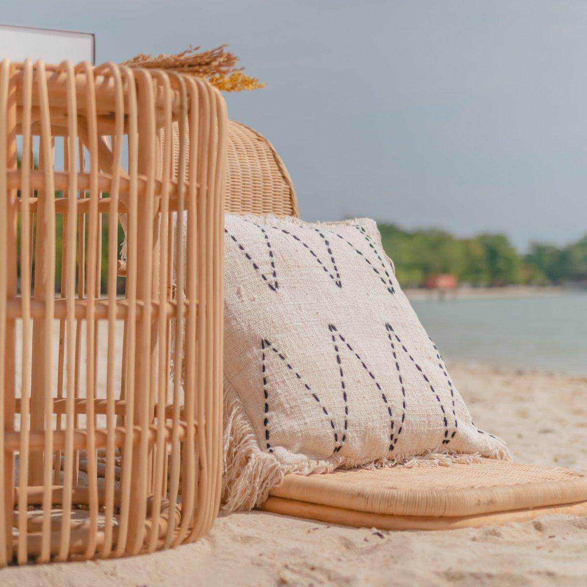 Rotan bijzettafel en kussen op strand, ideaal voor een stijlvolle, duurzame inrichting thuis of buiten.