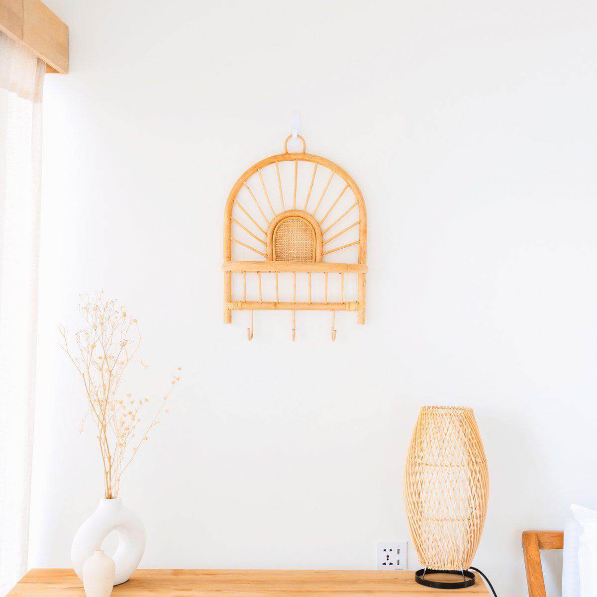 Rotan wandplank met boho-design en haken ingericht op een witte muur, naast een houten tafel en een stijlvolle lamp.
