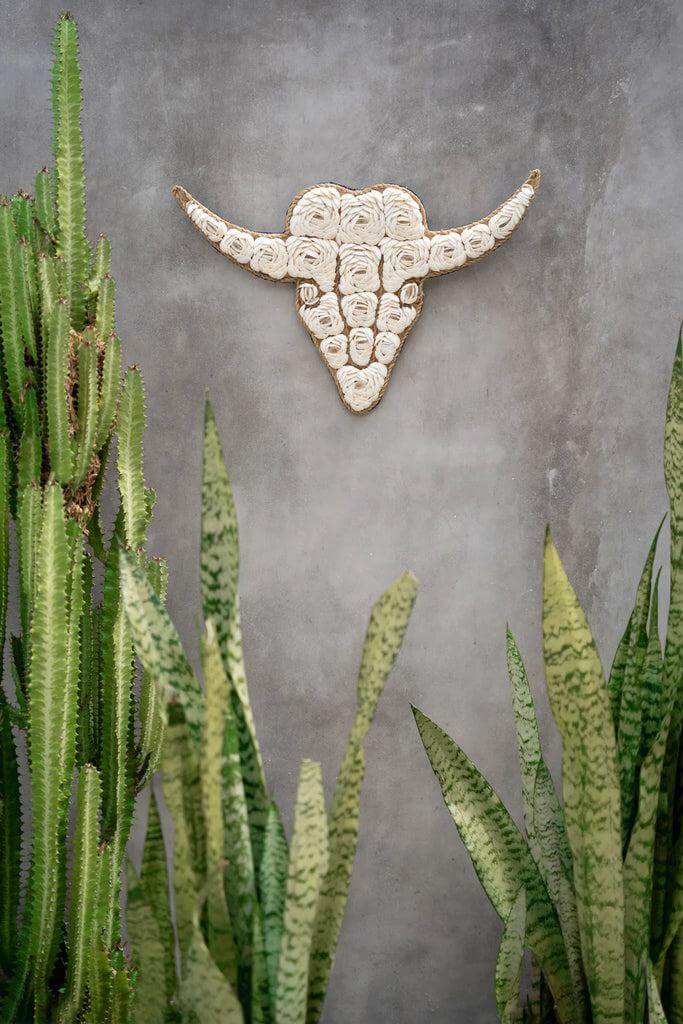 Witte decoratieve stier van schelpen in bloemmotief aan de muur tussen groene planten, een uniek interieuraccessoire.