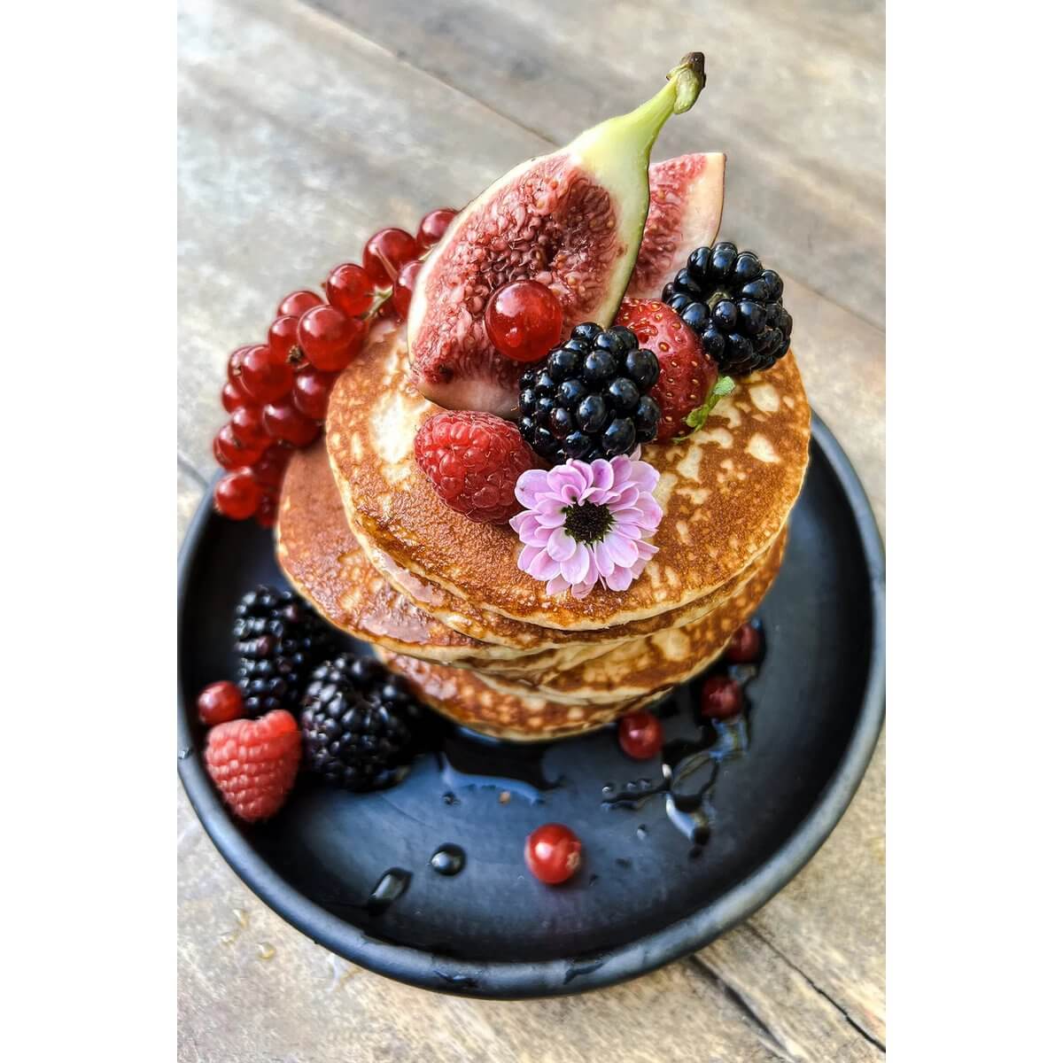 Stack of pancakes topped with figs, berries, and a flower on a black plate.