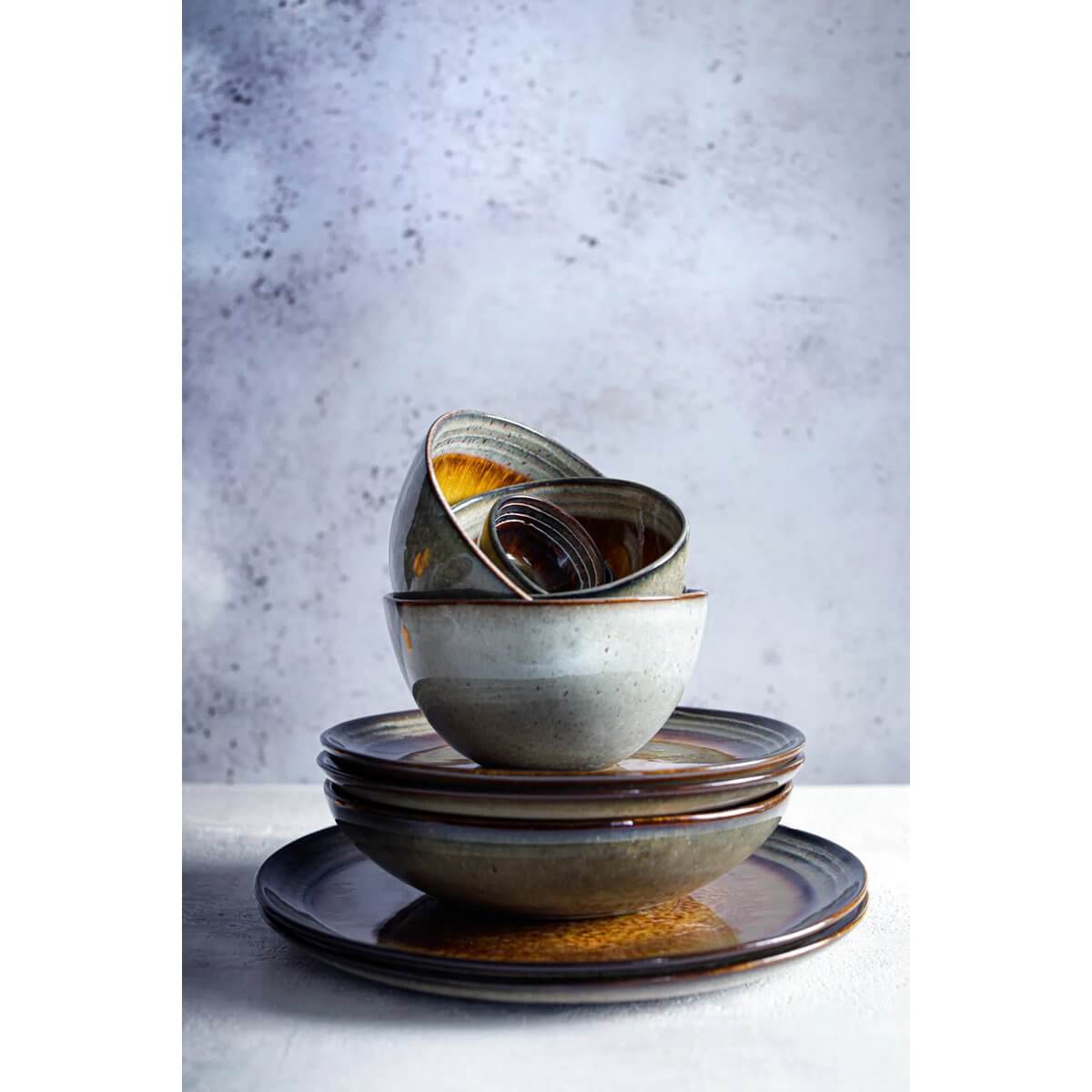 Stapeltjes keramische borden en kommen in aardetinten met variërende kleuren zoals bruin, groen en diepblauw, tegen een neutrale achtergrond.