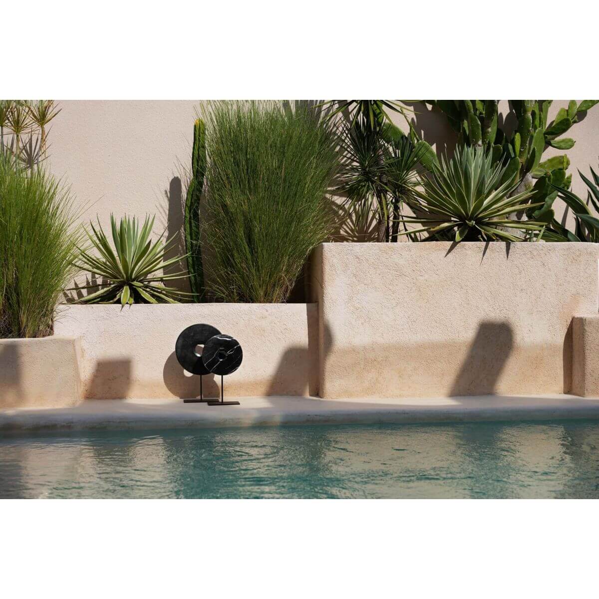 Zwarte marmeren schijf op standaard als stijlvol decor naast een zwembad, omringd door groene planten in een moderne buitenomgeving.