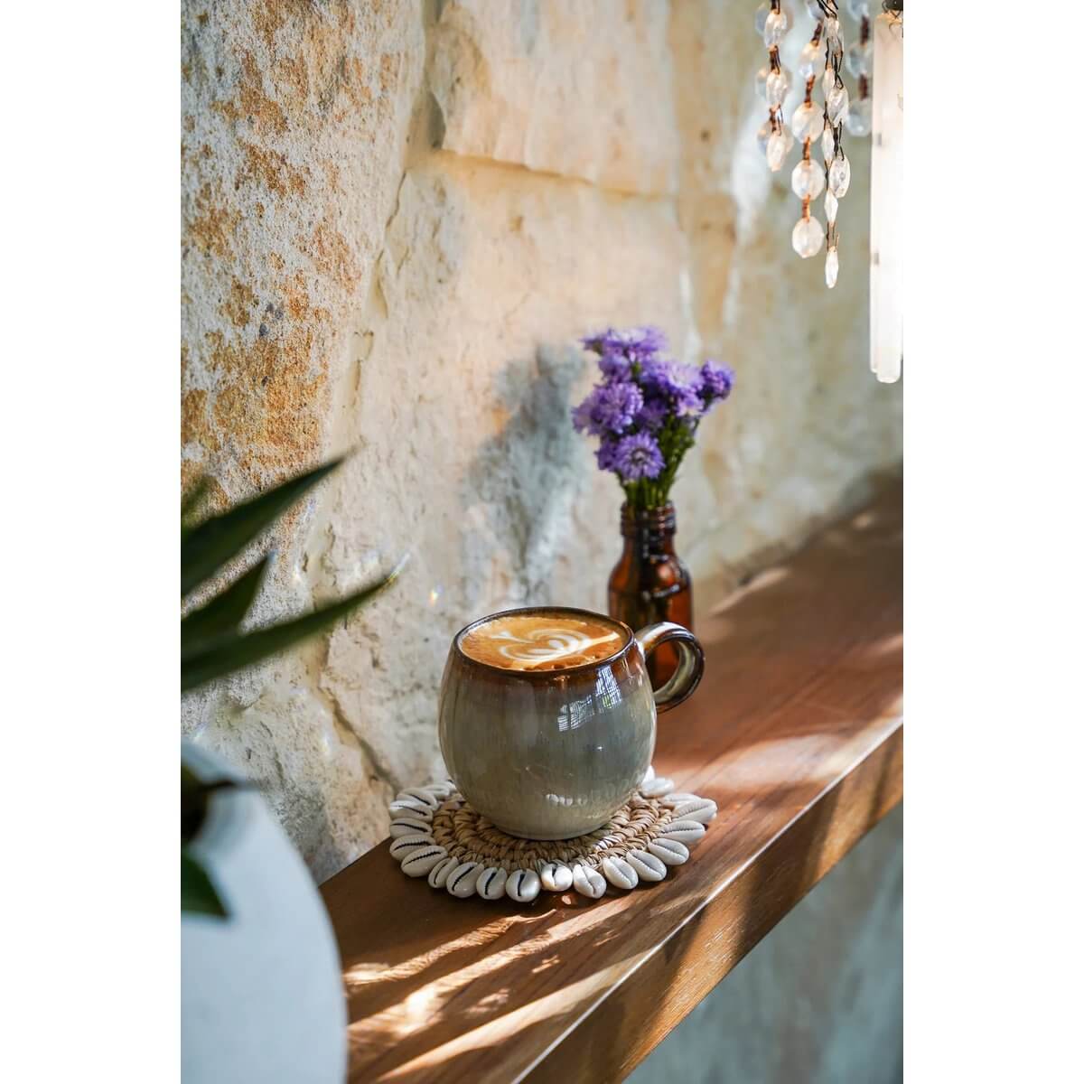 Bohemian decor: Raffia shell coaster with mug on a rustic table, adorned with cowrie shells, adding charm to a cozy nook.