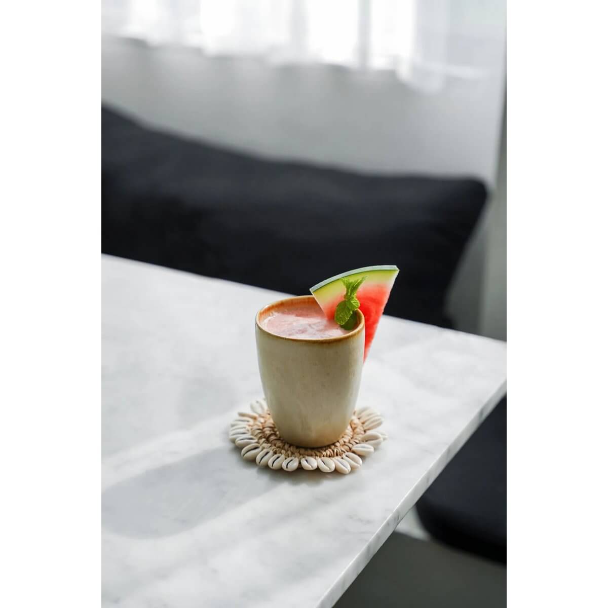 Bohemian Raffia Shell Coaster by Bazar Bizar Living on a table with a tropical drink and watermelon slice.