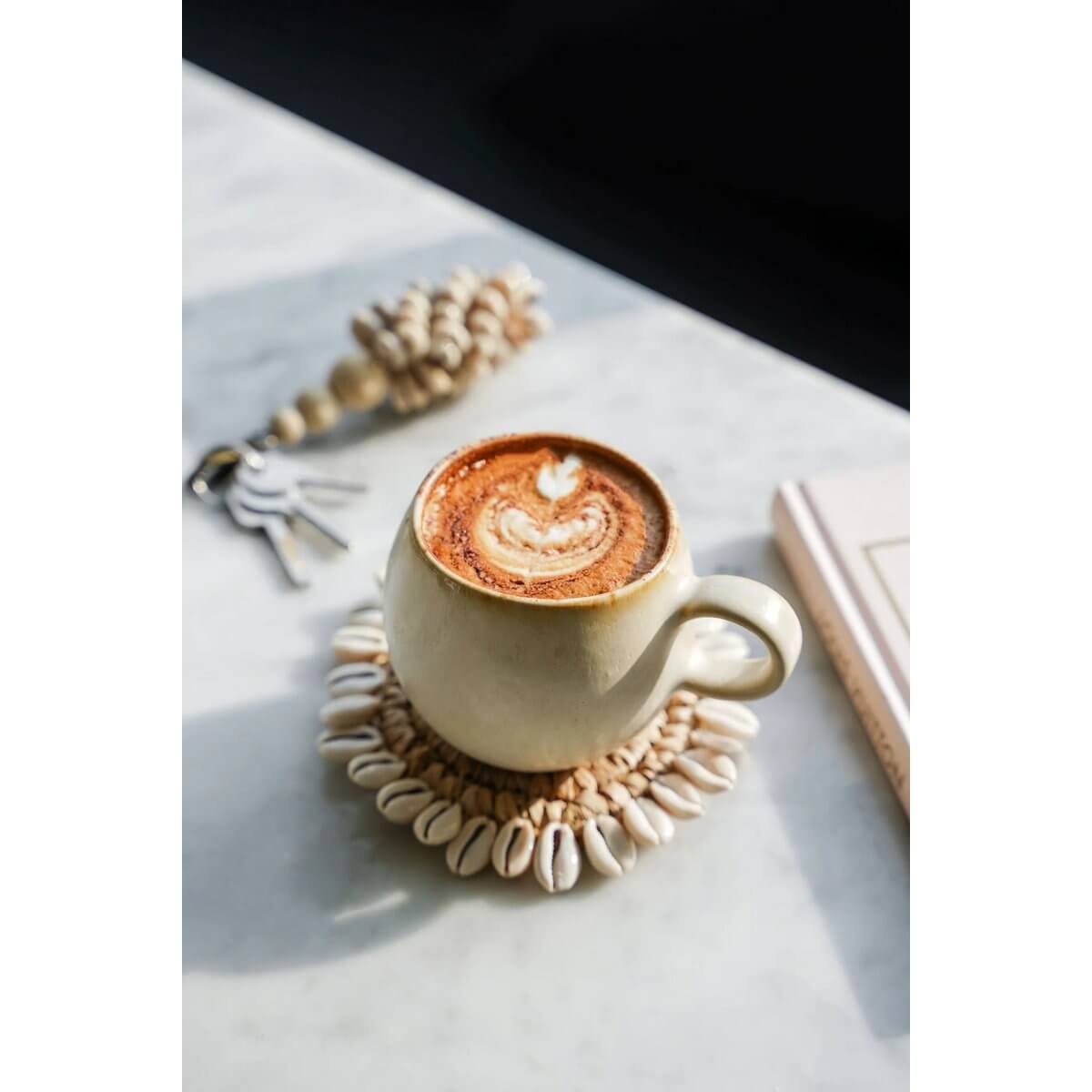 Bohemian Raffia Shell Coaster under a coffee cup on a marble table, decorated with kaurischelpen, enhancing Balinese vibes.