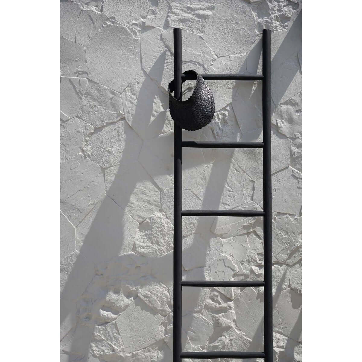Bohemian style black bamboo sunshade hat hanging on a black ladder against a textured white wall, perfect for spring and summer.