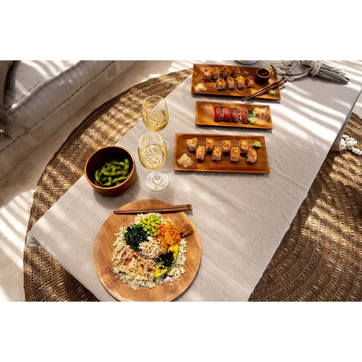 A Bohemian-style dining table setting with The Teak Root Round Plate, sushi, edamame, and wine glasses, creating a warm wooden ambiance.