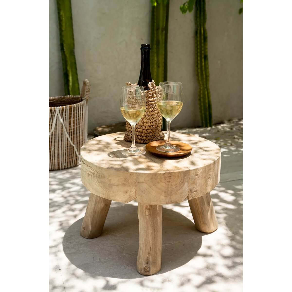 Rustic wooden table with wine glasses and a bottle under dappled sunlight, surrounded by lush greenery. Perfect bohemian setting.