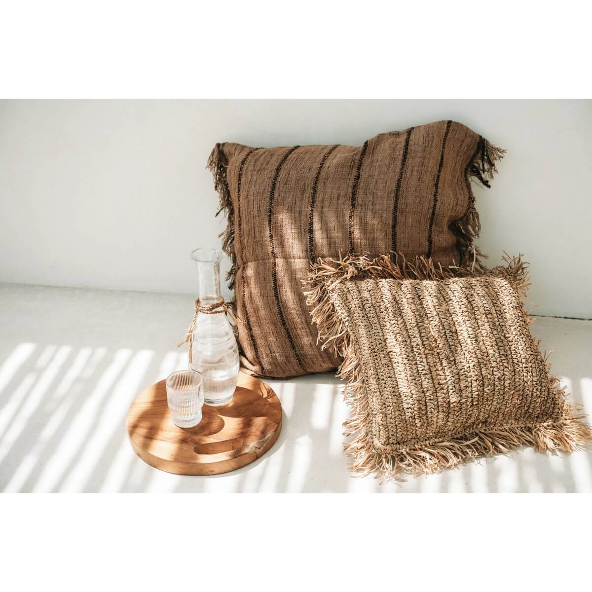 Twee bruine kussens met franjes, gelegen naast glazen flessen op een ronde teakhouten schaal op een zonnige achtergrond.