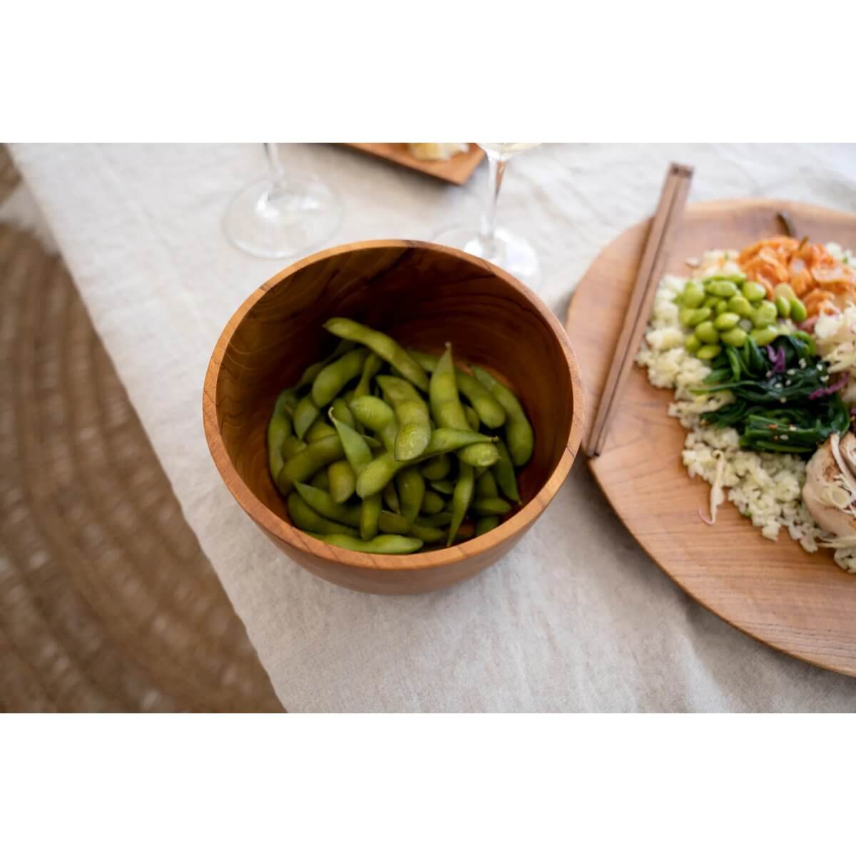 Bohemian dining setup with a teak salad bowl filled with edamame, highlighting sustainable style and elegance on a woven mat.