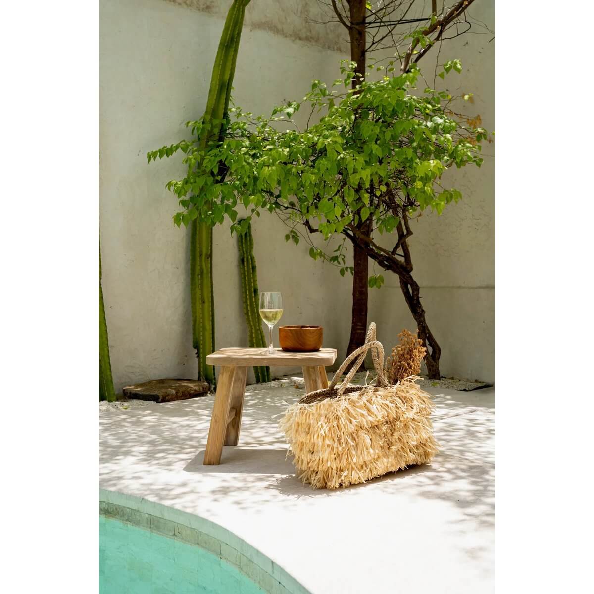 Bohemian outdoor setup with a teak Wortel Salade Bowl on a table, next to a straw bag and glass of wine by the pool.