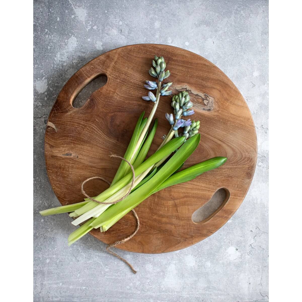 Ronde teakhouten snijplank dienblad met handvatten, versierd met verse bloemen en groen op een grijze achtergrond.