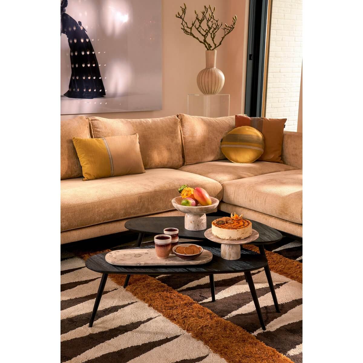 Stijlvol interieur met Finnos taartplateau in beige marmer op een zwarte salontafel, omgeven door luxe beige bank en kleurrijke kussens.