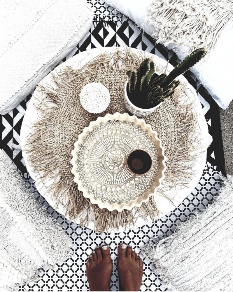 Raffia placemat met kauri schelpen op tropische tafelsetting, omgeven door kussens en een cactusplant, voor eilandstijl interieur.