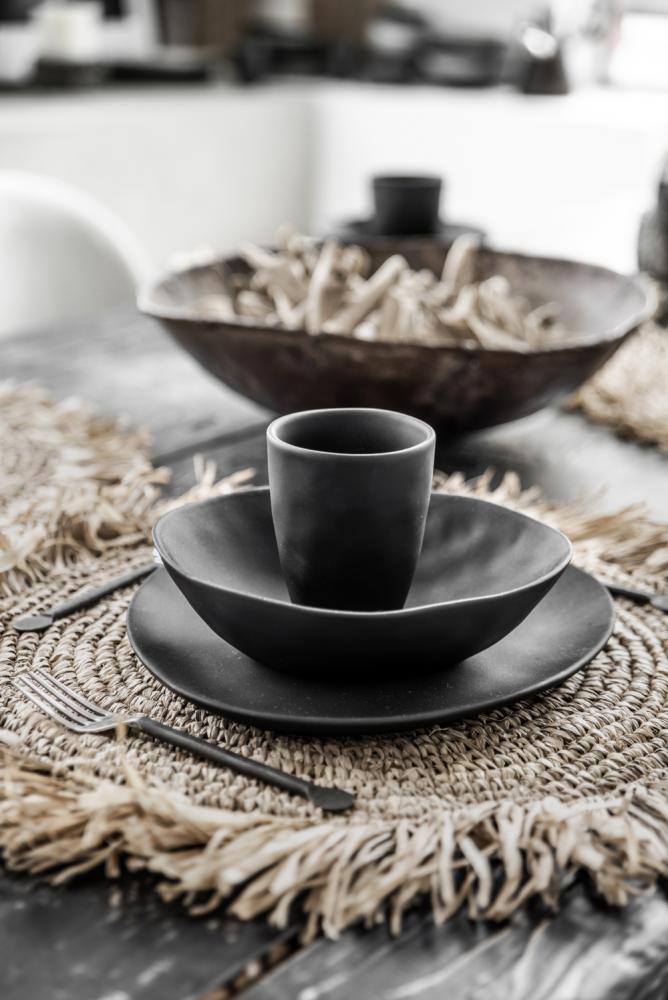 Elegante tafelsetting met handgeweven raffia placemat en zwart serviesgoed voor een tropische sfeer.