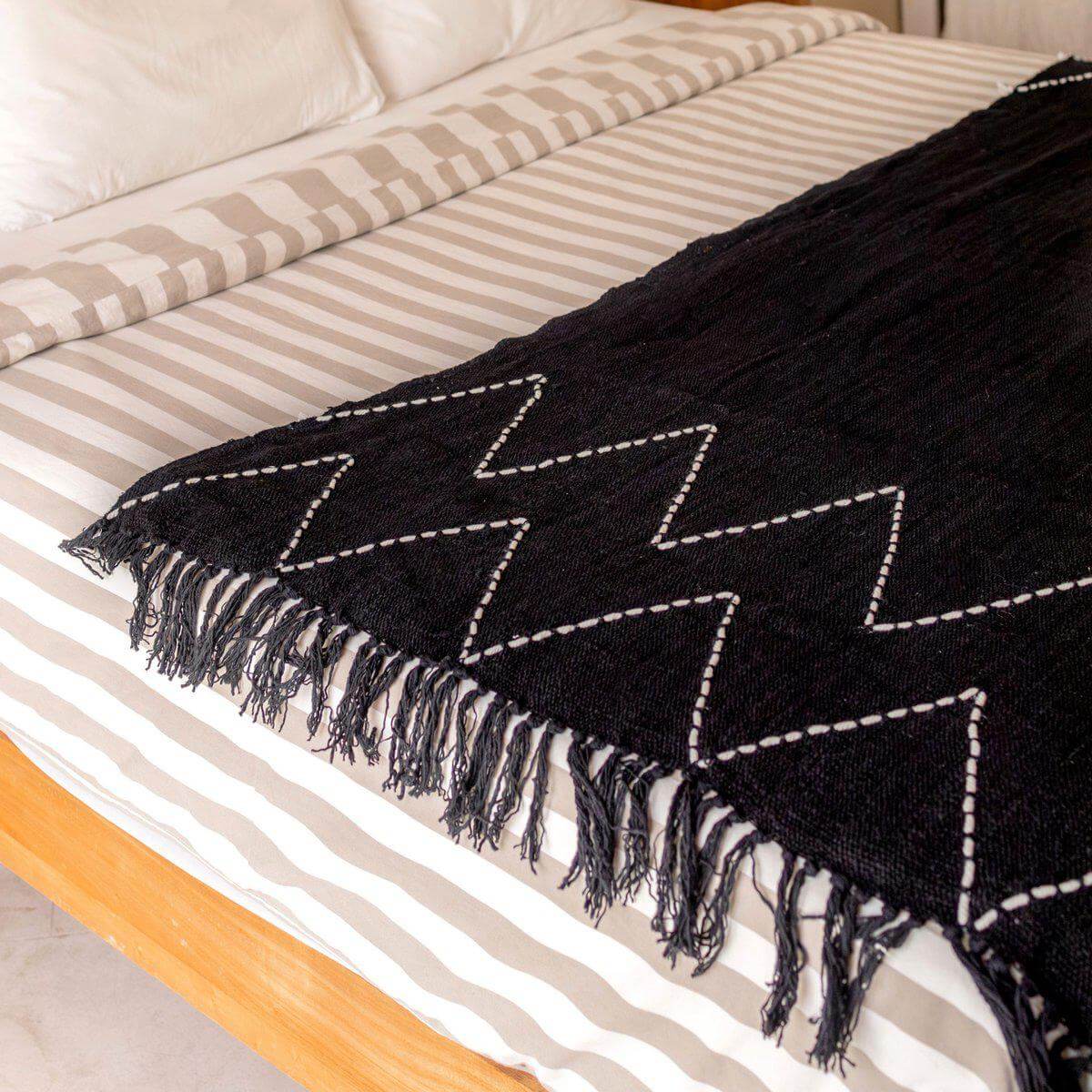 Zwarte handgeweven katoenen deken 140x200 cm op bed, boho stijl met zigzag patroon, duurzaam en plasticvrij verpakt.