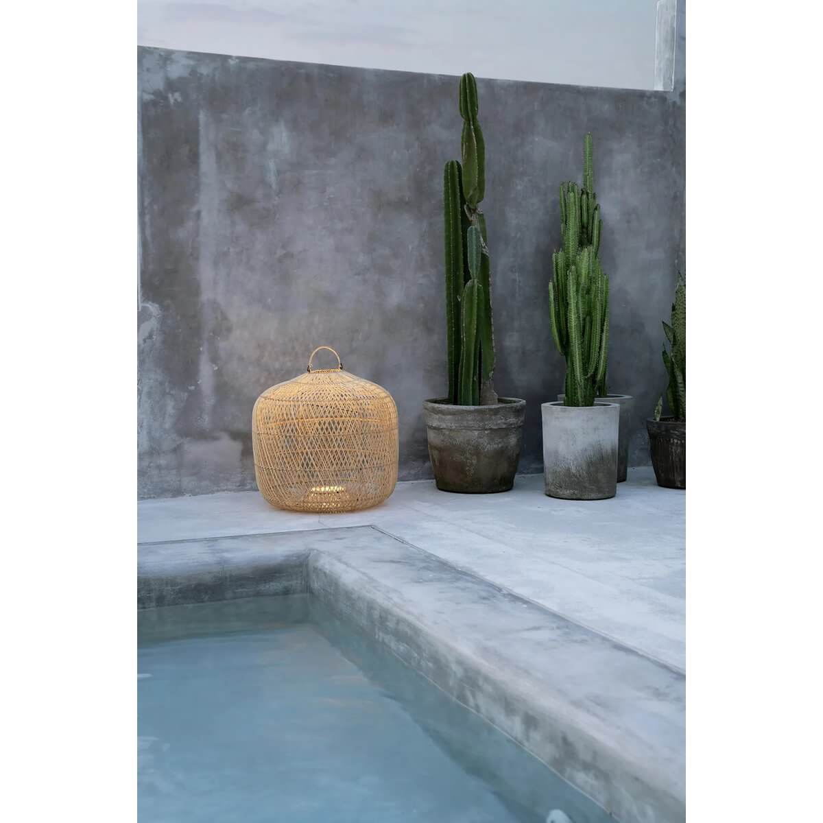 Bohemian De Batu Bolong rattan lamp, natural color, placed by indoor cactus plants, near a modern poolside setting.