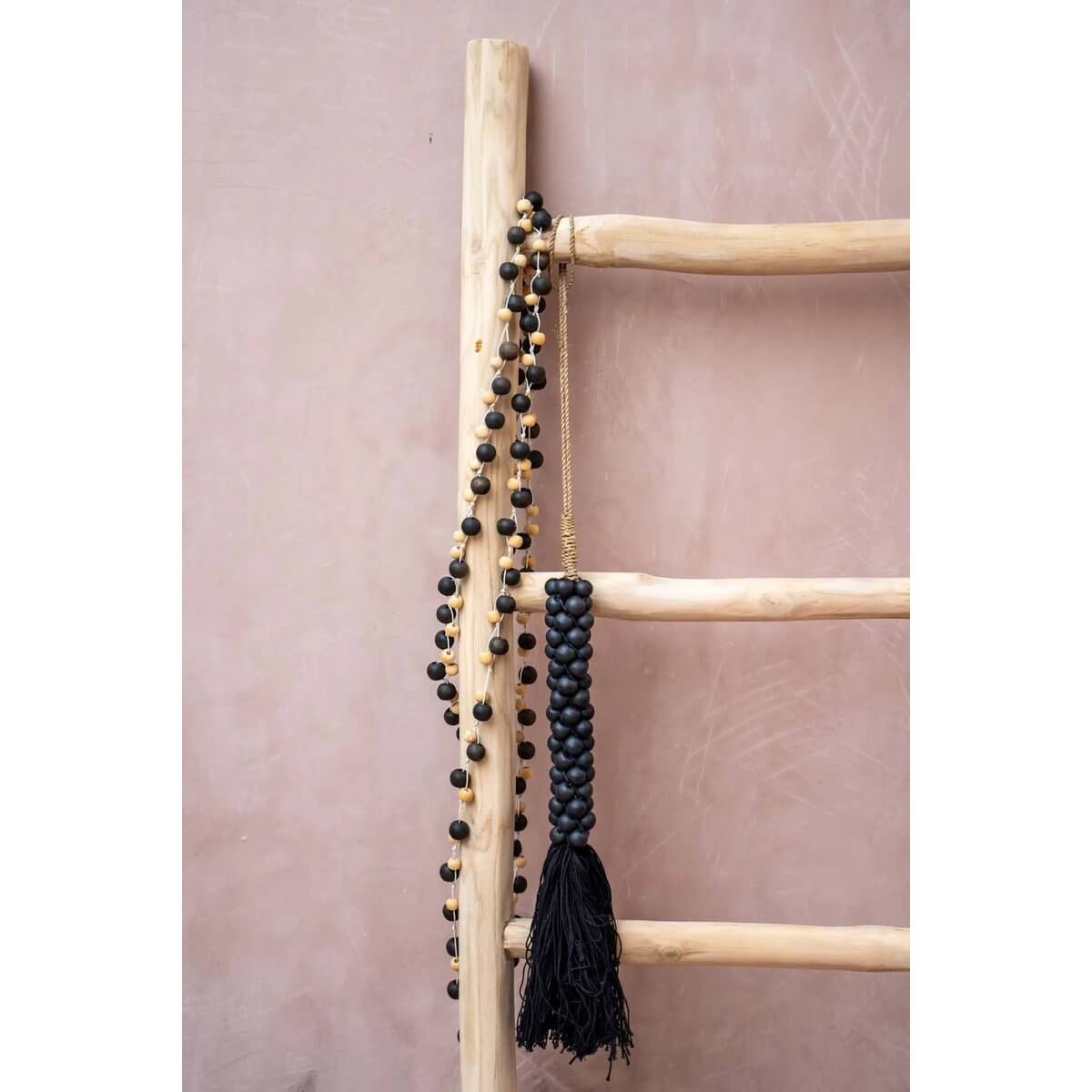 Bohemian De Canggu ketting in natuurlijk zwart hout stijlvol gedrapeerd over een houten ladder tegen een roze achtergrond.