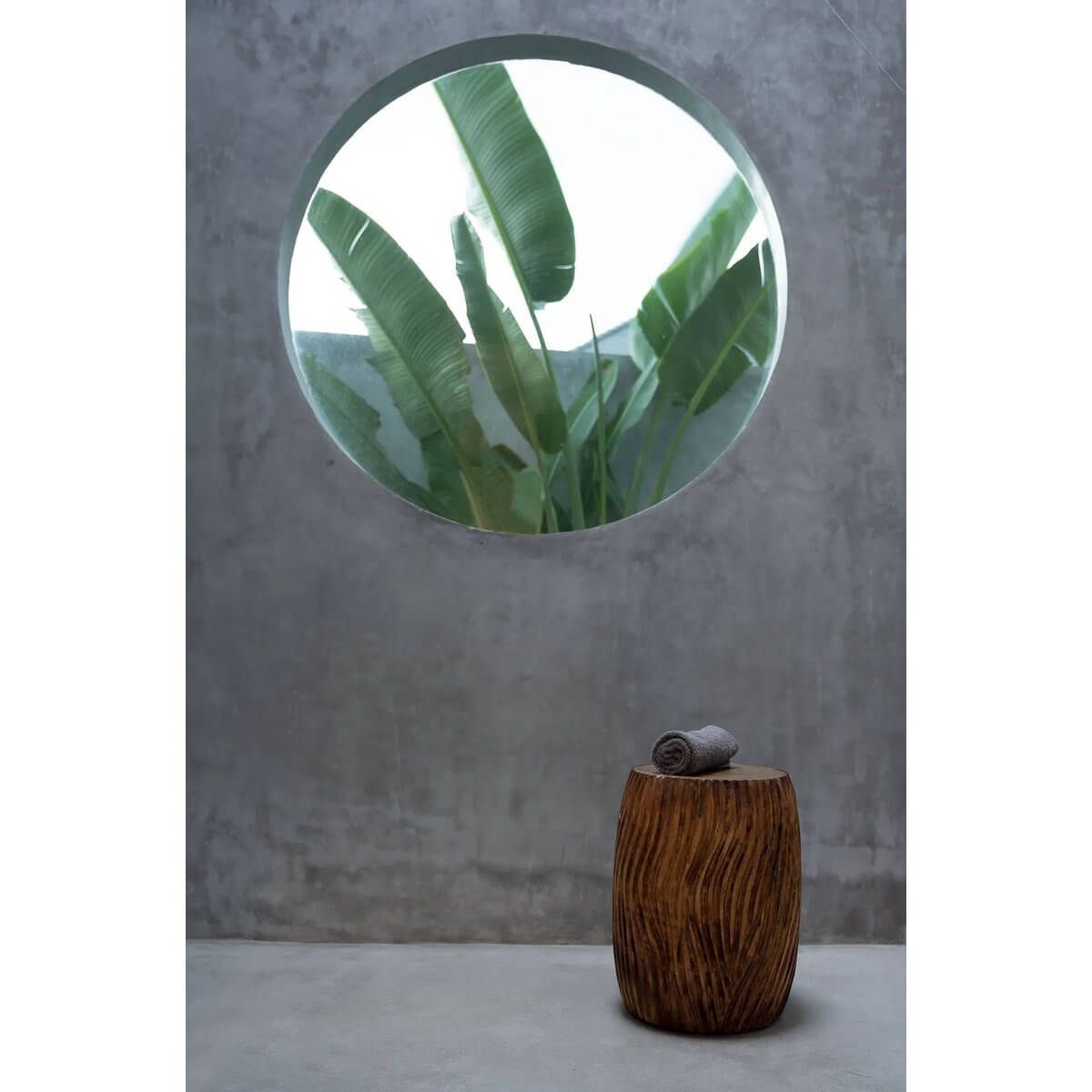 Bohemian style De Celebes Stool in natural suar wood with carved design, placed under a round window with leafy view.