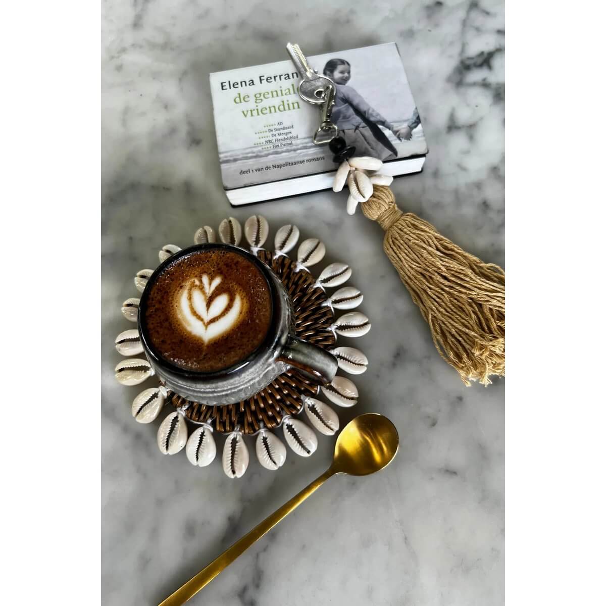 Bohemian Cowrie Tassel Keychain in Mocca next to coffee on shell coaster and book on marble surface.