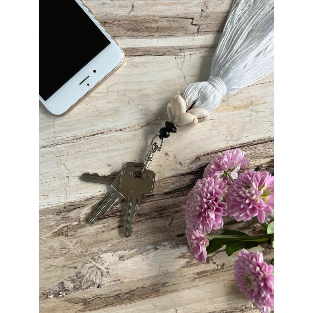 Bohemian Cowrie Tassel Keychain with keys on a rustic surface, next to pink flowers and smartphone.