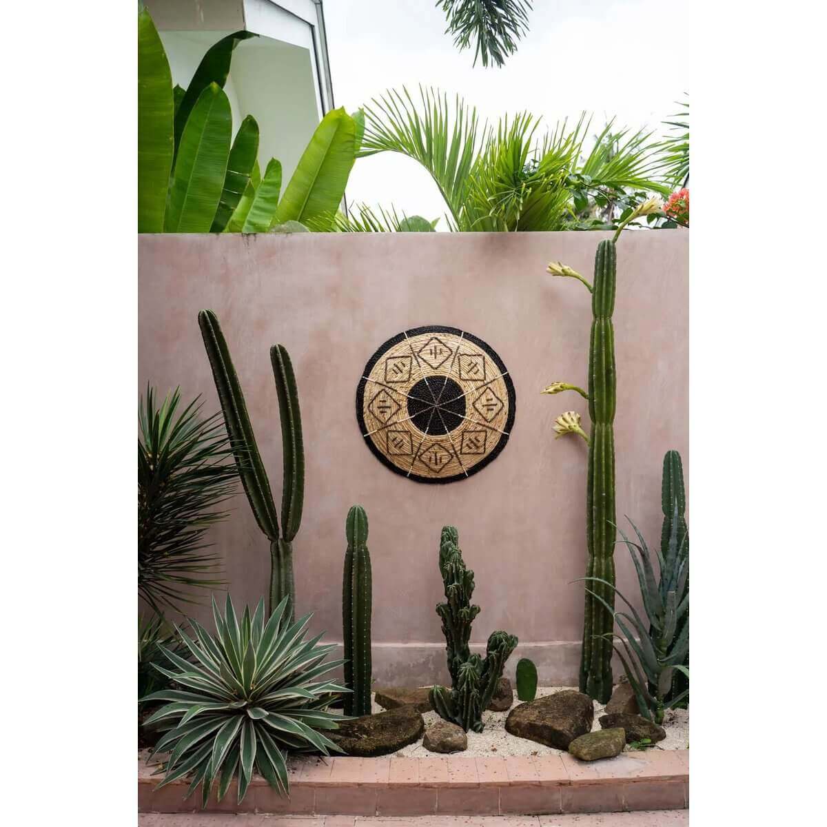 Handgeweven bamboe wandbord met zwarte grafische details als etnische decoratie tegen een muur, omgeven door cactussen.