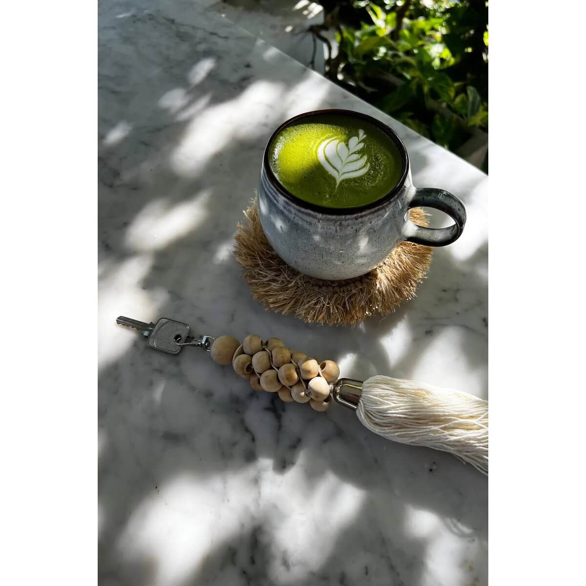 Bohemian keychain with natural white wooden beads and cotton tassel beside a matcha latte on a marble table.