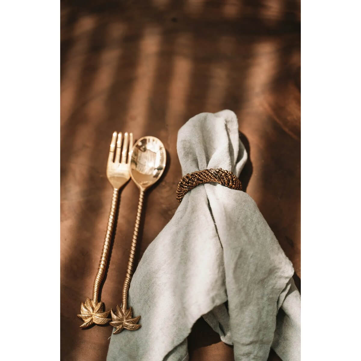 Bohemian style gold beaded napkin ring on a linen napkin, next to elegant golden cutlery on a rustic table.