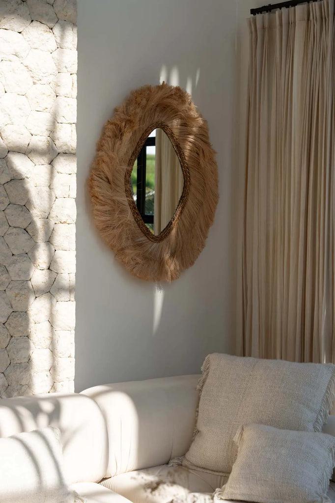 De La Duchessa Naturel Spiegel aan de muur, vangt zonlicht in een moderne woonkamerinterieur met witte bank.