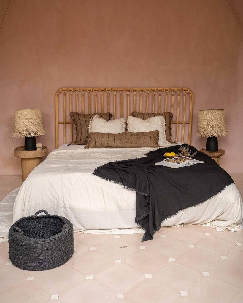 Stijlvolle slaapkamer met rotan hoofdsteun, beige en zwarte kussens en dekens, en rustieke tafellampen voor een boho sfeer.
