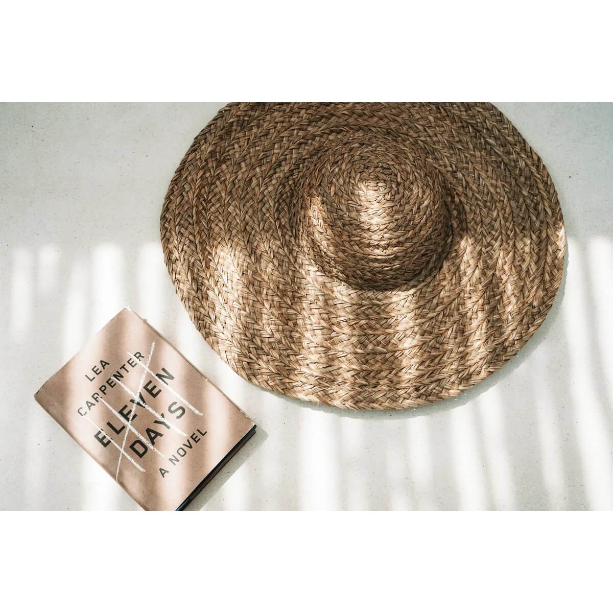 Bohemian De Playa Hat made of waterhyacinth next to a novel, casting shadows in sunlight. Perfect summer accessory for cozy days outdoors.