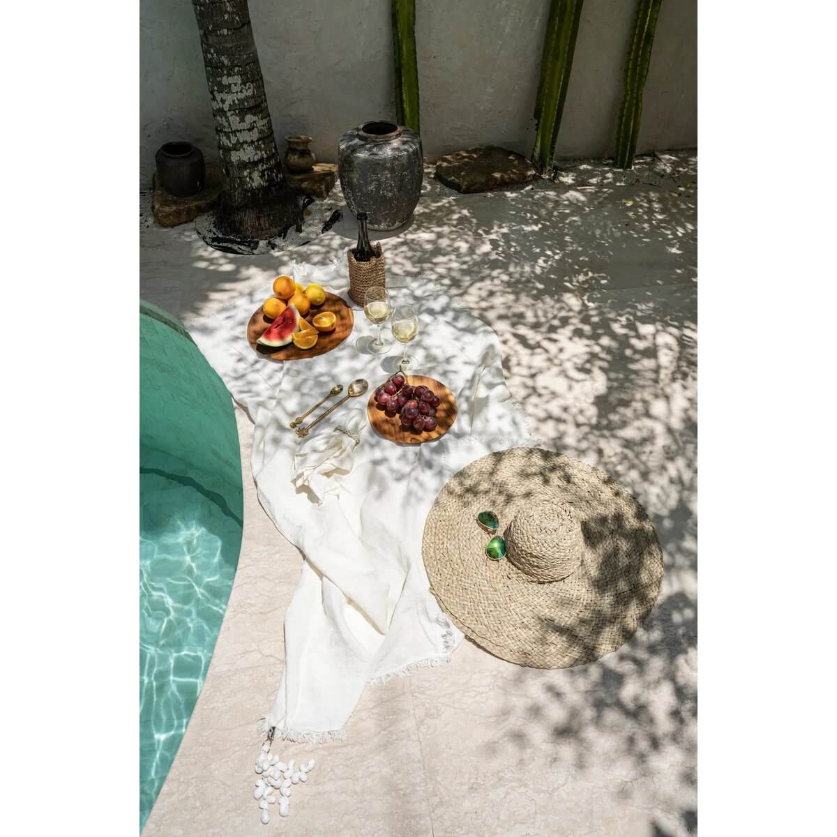 Tropical poolside scene with a beige De Playa bohemian hat on a white cloth, surrounded by fresh fruits and shaded by palm trees.