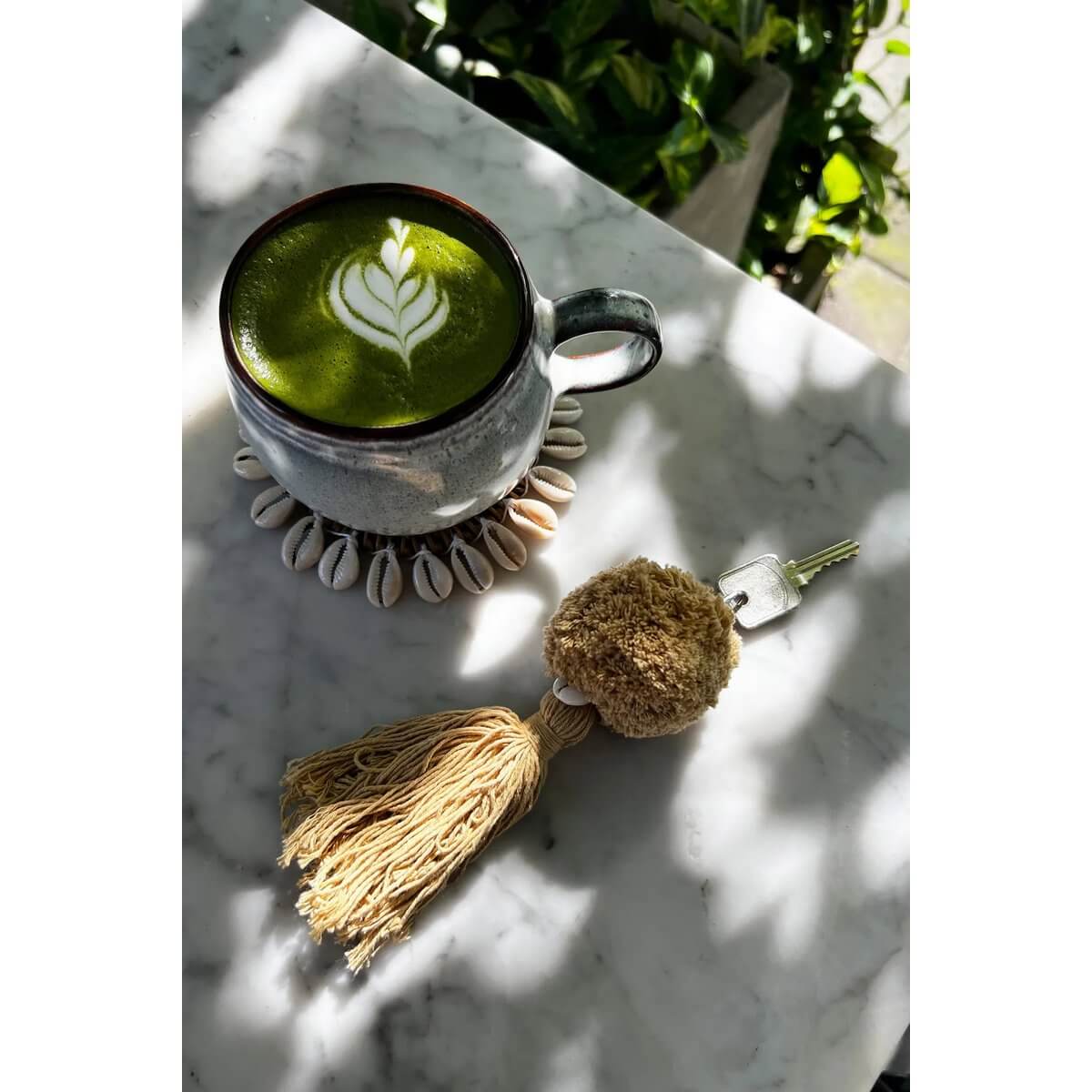 Cozy bohemian scene with a mokka pompom keychain on marble, next to a green matcha latte in a ceramic cup.
