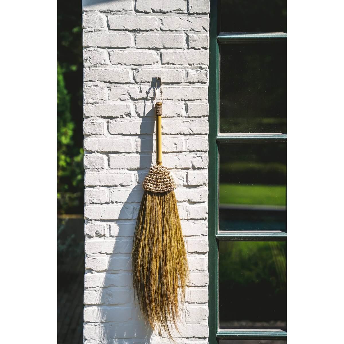 Handmade Indonesian bohemian broom with natural fibers, decorative hanging on white brick wall. Ideal for interior or covered areas.