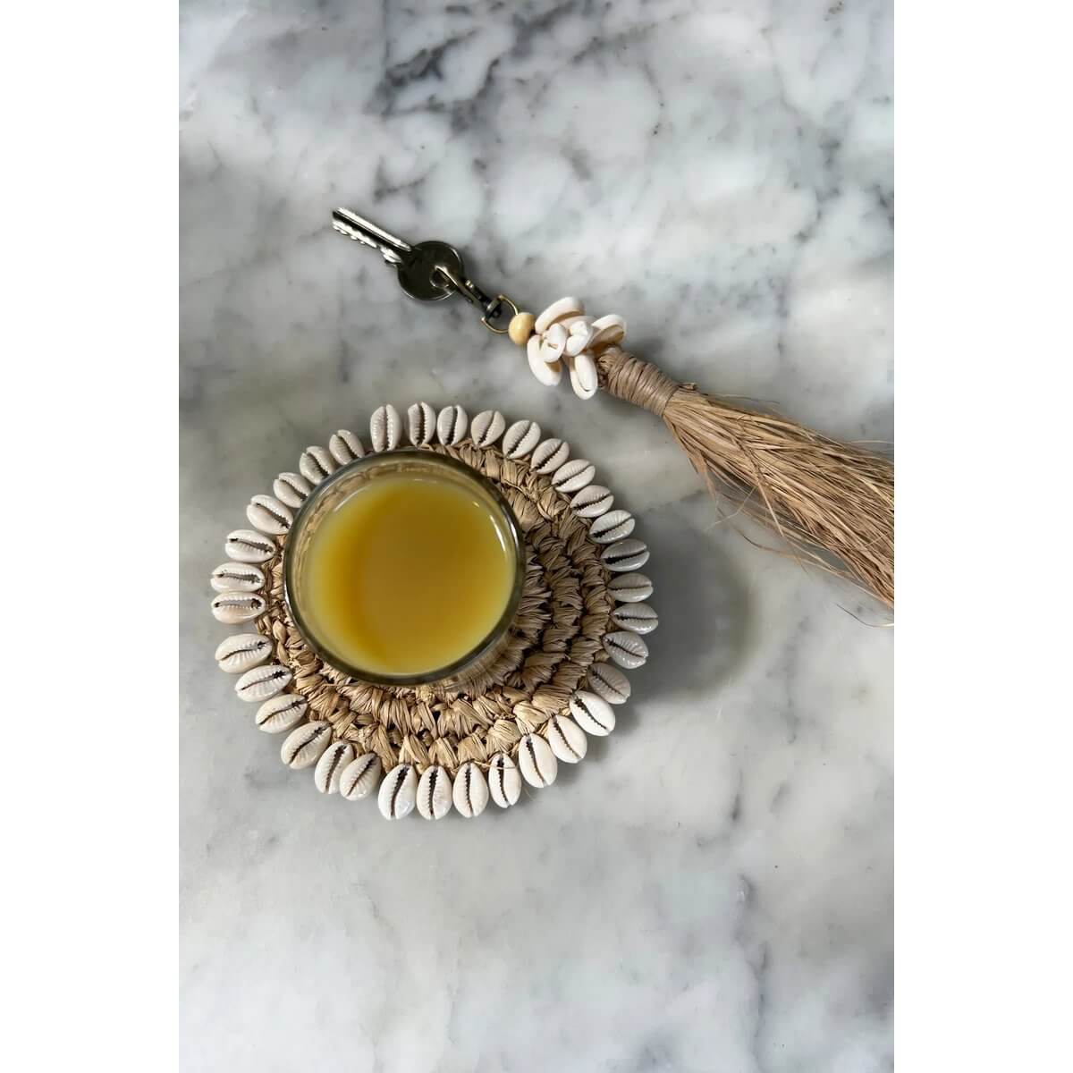 Bohemian De Raffia Kauri-schelp sleutelhanger on marble surface beside shell coaster with drink.