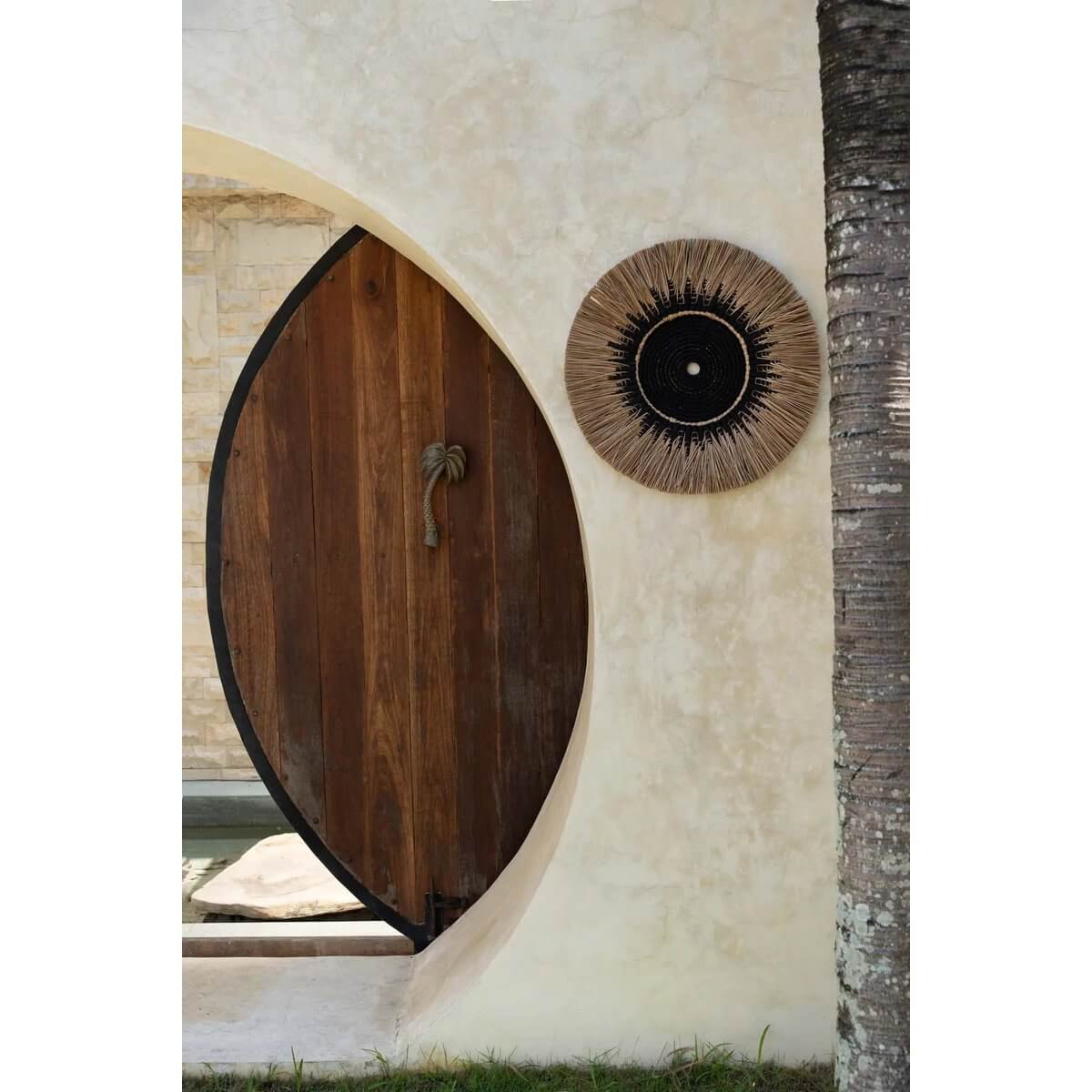 De Raffia Macramé Muurdeco in zwart en naturel, handgemaakt van katoen en raffia, hangt naast een houten deur in een boheemse stijl.