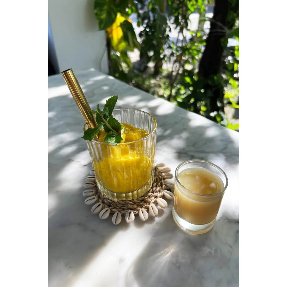Bohemian De Seagrass Shell Coaster with glasses of fresh juice on a sunlit table, surrounded by lush greenery.