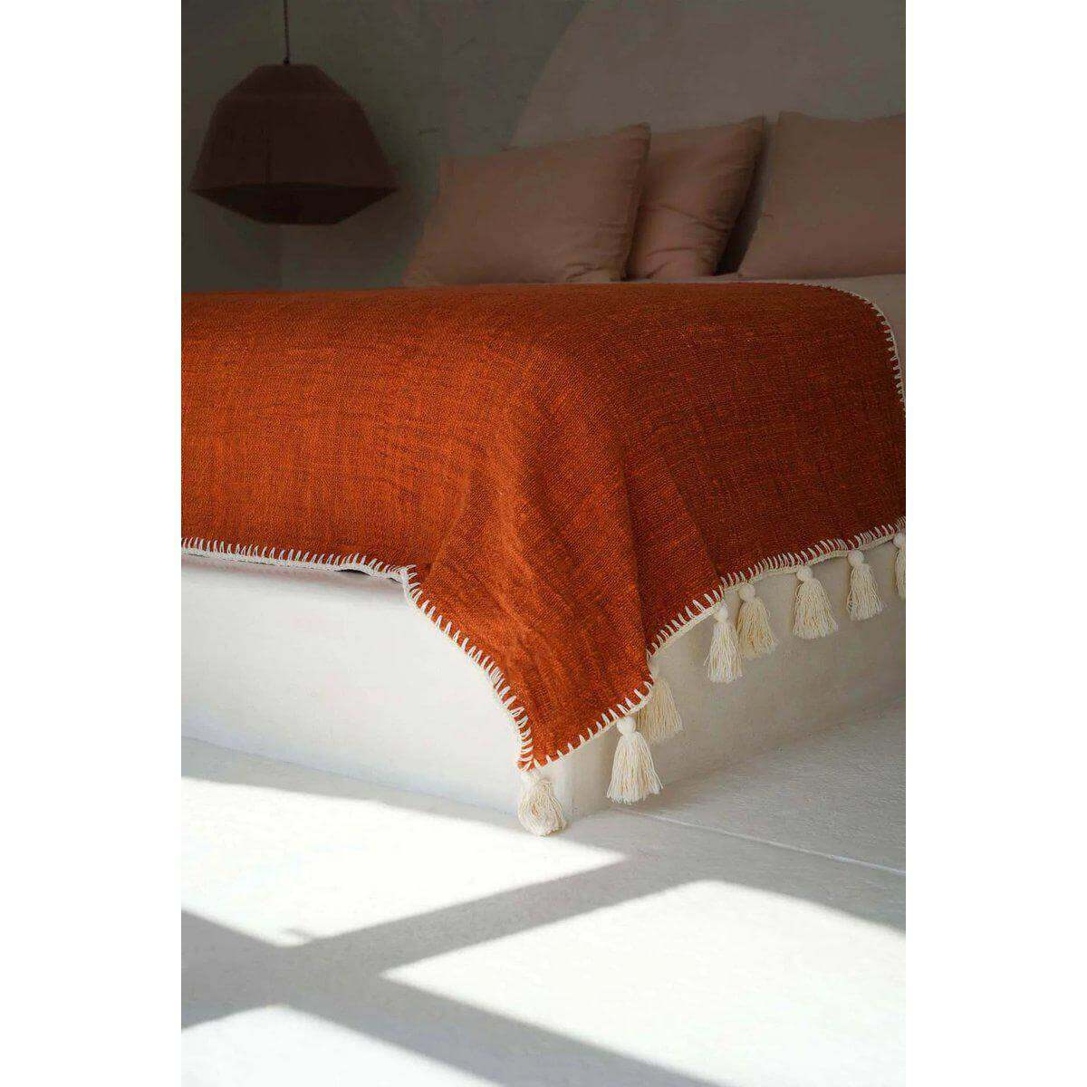 De Stitch Bedsprei in terracotta, stijlvol gedrapeerd over een bed met kwastjes, voegt een warme en gezellige sfeer toe aan de slaapkamer.