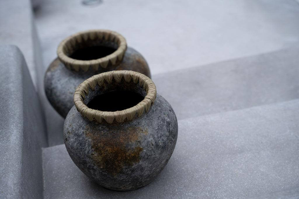Twee antiek grijze terracotta vazen met raffiadetail, ideaal voor droogbloemen, gepositioneerd op betonnen trappen.