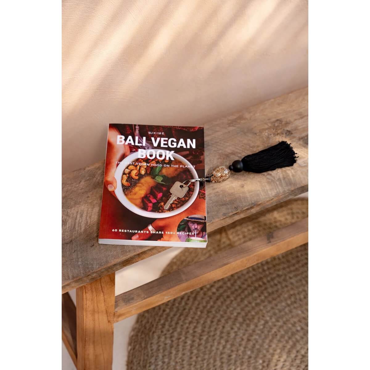 Bali Vegan Book on a wooden bench beside a black tassel keychain, showcasing a rustic and bohemian vibe.