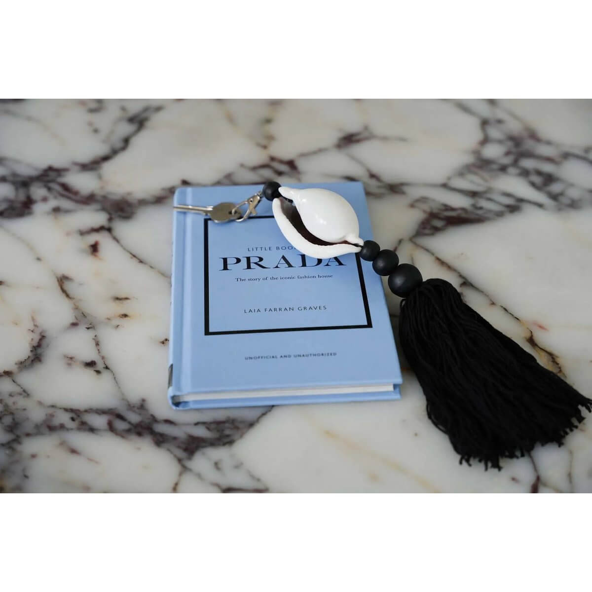 De Togian sleutelhanger in zwart-wit, houten en katoenen materialen, met een witte schaal, ligt op een blauw boek voor een bohemian touch.