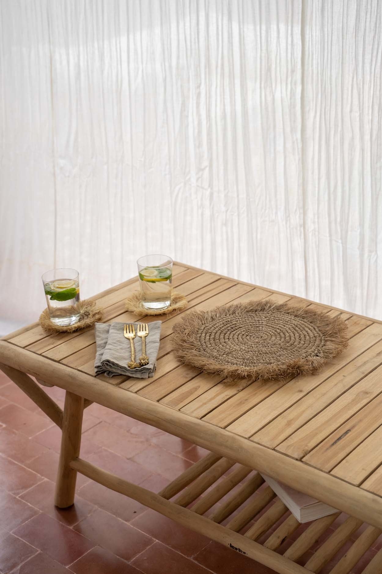De Tulum salontafel van teakhout met zomerse uitstraling, ideaal voor terras en tuin, met drankjes en placemat erop.