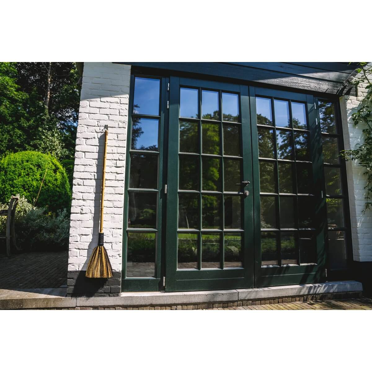 Bohemian decorative broom made of Indonesian grass leaning against a stylish black glass door in a serene garden setting.