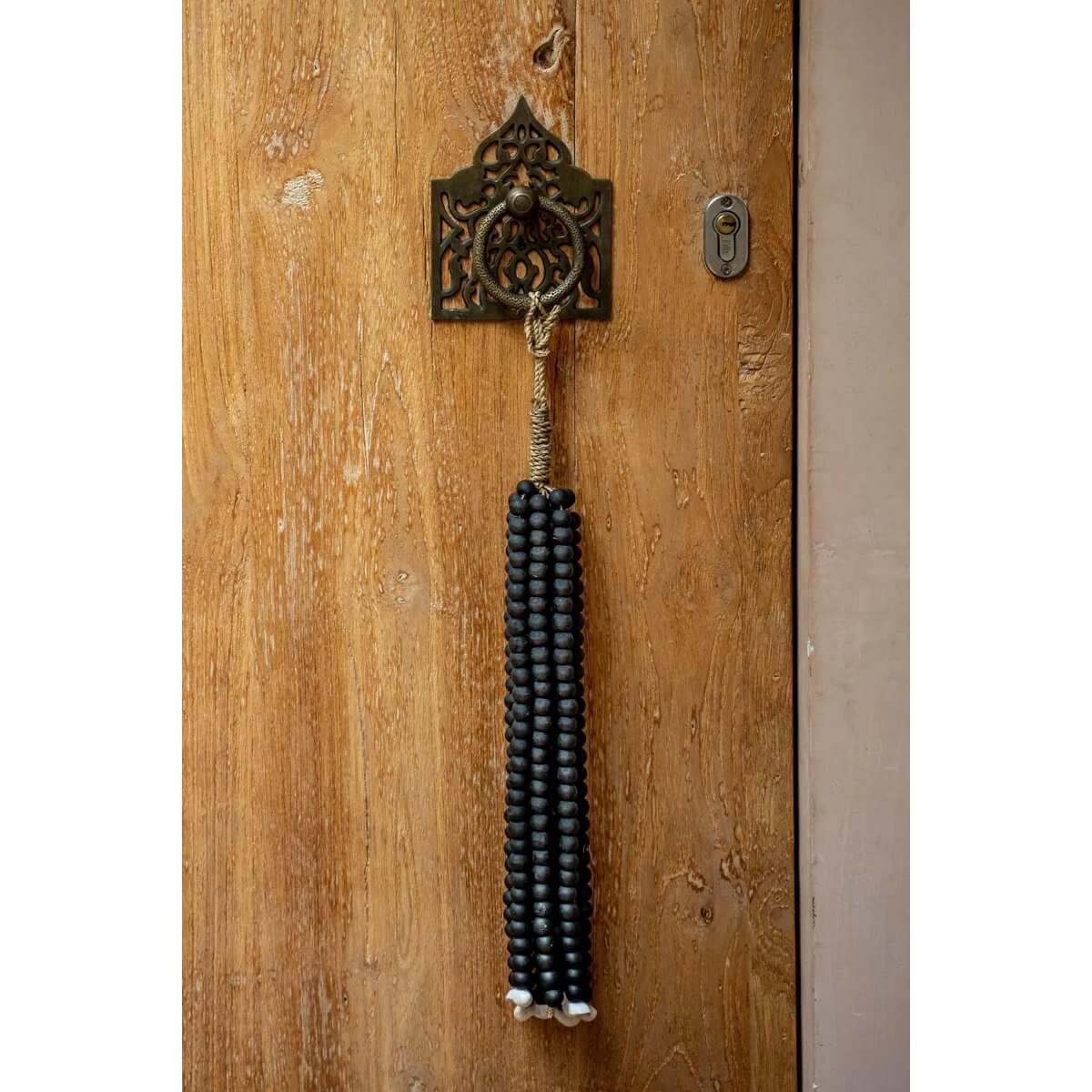 Black wooden bead tassel by Bazar Bizar, handcrafted by Balinese artisans, hanging on a wooden door for bohemian home decor.