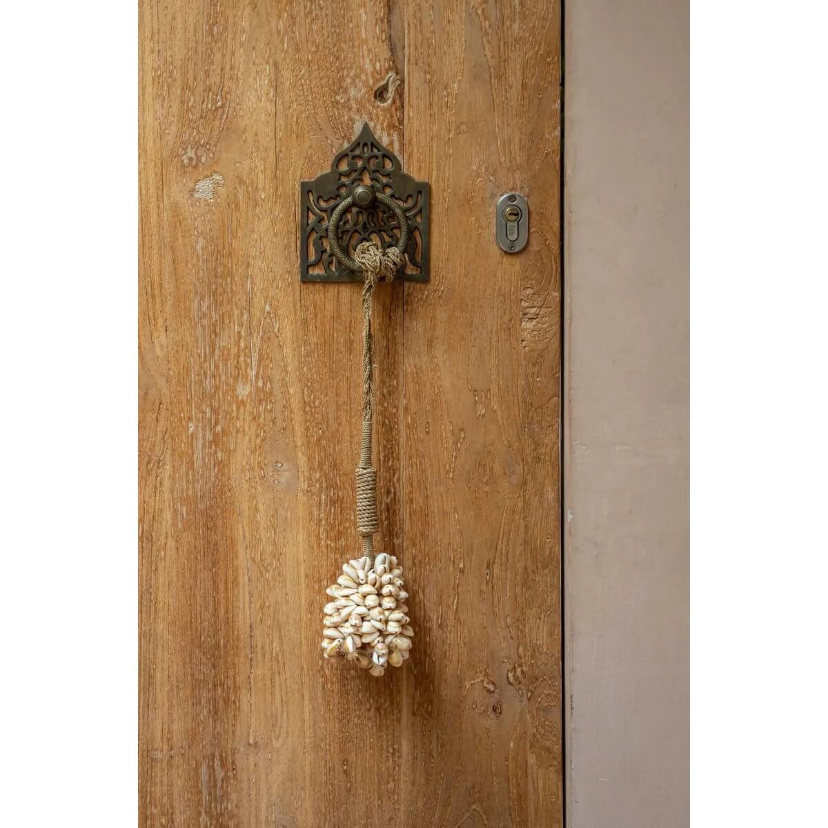Handmade bohemian pink shell tassel hanging on a rustic wooden door, adding exotic coastal charm to home decor. By Bazar Bizar Living.