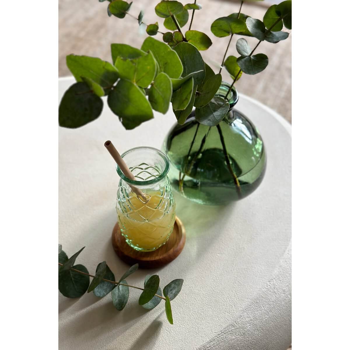 Tropisch bohemian tafelsetting met een blauw glazen ananasbeker gevuld met sap en groene bladeren in een vaas.